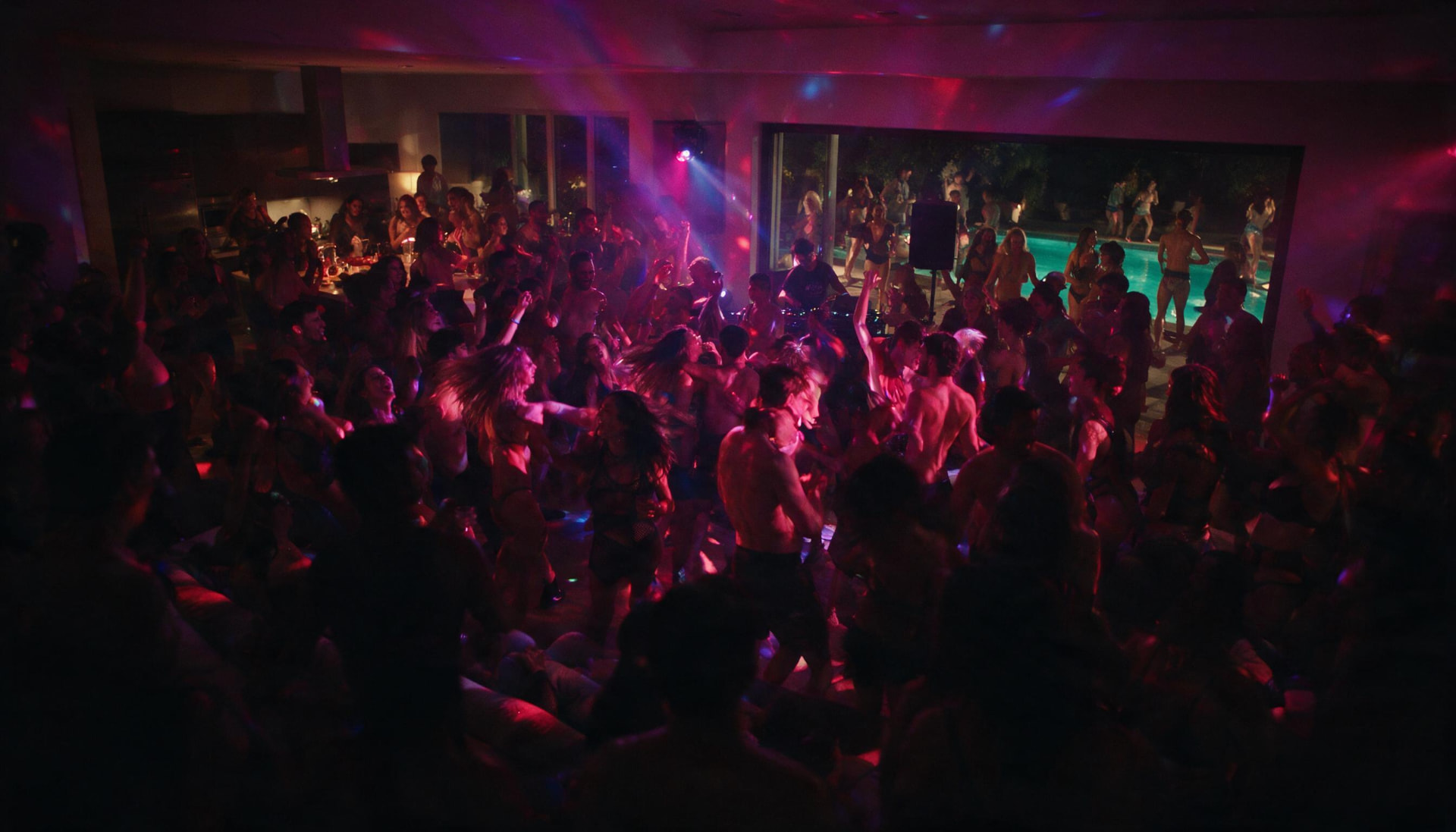 Packed private house party dance floor under deep pink lighting with the pool visible outside